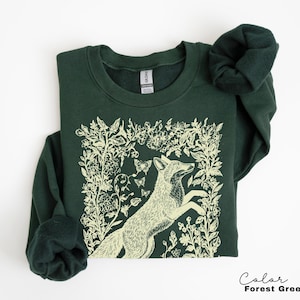 Pu&ograve; includere: Felpa verde foresta con un'illustrazione color crema di una volpe che salta tra accenti floreali e farfalle. Il testo "Color Forest Green" &egrave; in basso a destra.
