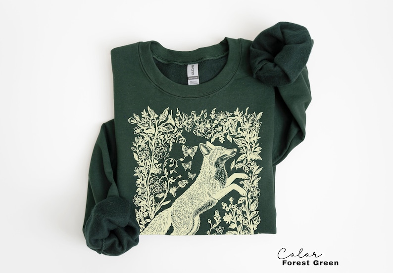 Pu&ograve; includere: Felpa verde foresta con un'illustrazione color crema di una volpe che salta tra accenti floreali e farfalle. Il testo "Color Forest Green" &egrave; in basso a destra.