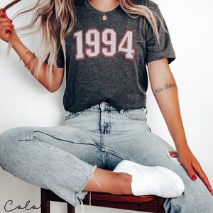 May include: A dark grey heather t-shirt with the year 1994 printed in white with a red outline. The shirt is being worn by a person with light blue jeans and white sneakers.