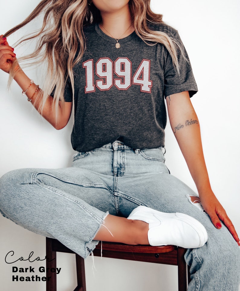 May include: A dark grey heather t-shirt with the year 1994 printed in white with a red outline. The shirt is being worn by a person with light blue jeans and white sneakers.