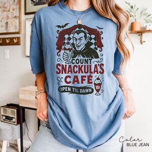 May include: Blue jean colored t-shirt featuring a graphic of Count Snackula's Cafe. The design includes a cartoon vampire holding a milkshake, bats, and the text "Count Snackula's Cafe, Open Til Dawn".