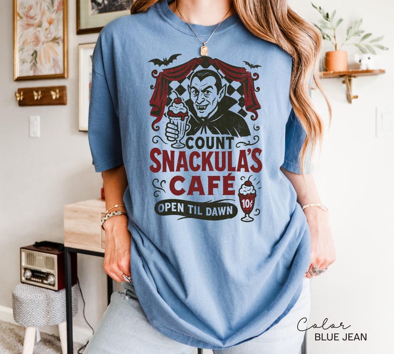 May include: Blue jean colored t-shirt featuring a graphic of Count Snackula's Cafe. The design includes a cartoon vampire holding a milkshake, bats, and the text "Count Snackula's Cafe, Open Til Dawn".