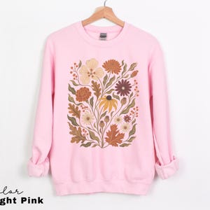Puede incluir: Una sudadera de cuello redondo en color rosa claro con un estampado gr&aacute;fico central de varias flores y hojas en tonos tierra apagados, incluyendo naranja, amarillo, marr&oacute;n y verde oliva. La prenda de manga larga tiene cuello, pu&ntilde;os y dobladillo acanalados.