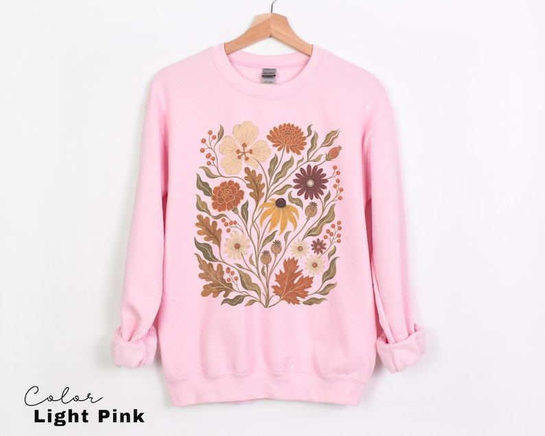 Puede incluir: Una sudadera de cuello redondo en color rosa claro con un estampado gr&aacute;fico central de varias flores y hojas en tonos tierra apagados, incluyendo naranja, amarillo, marr&oacute;n y verde oliva. La prenda de manga larga tiene cuello, pu&ntilde;os y dobladillo acanalados.