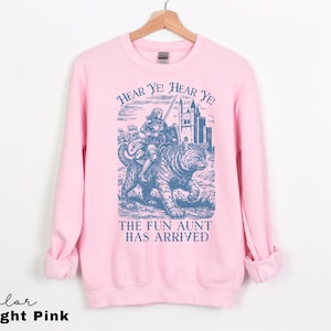 May include: Light pink sweatshirt with a blue graphic of a person riding a tiger, with the text "Hear Ye! Hear Ye! The Fun Aunt Has Arrived". The sweatshirt is hanging on a wooden hanger.