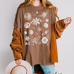 Puede incluir: Una camiseta marr&oacute;n con un estampado gr&aacute;fico blanco de planetas, estrellas y flores. El texto "Color Espresso" est&aacute; impreso debajo de la imagen.