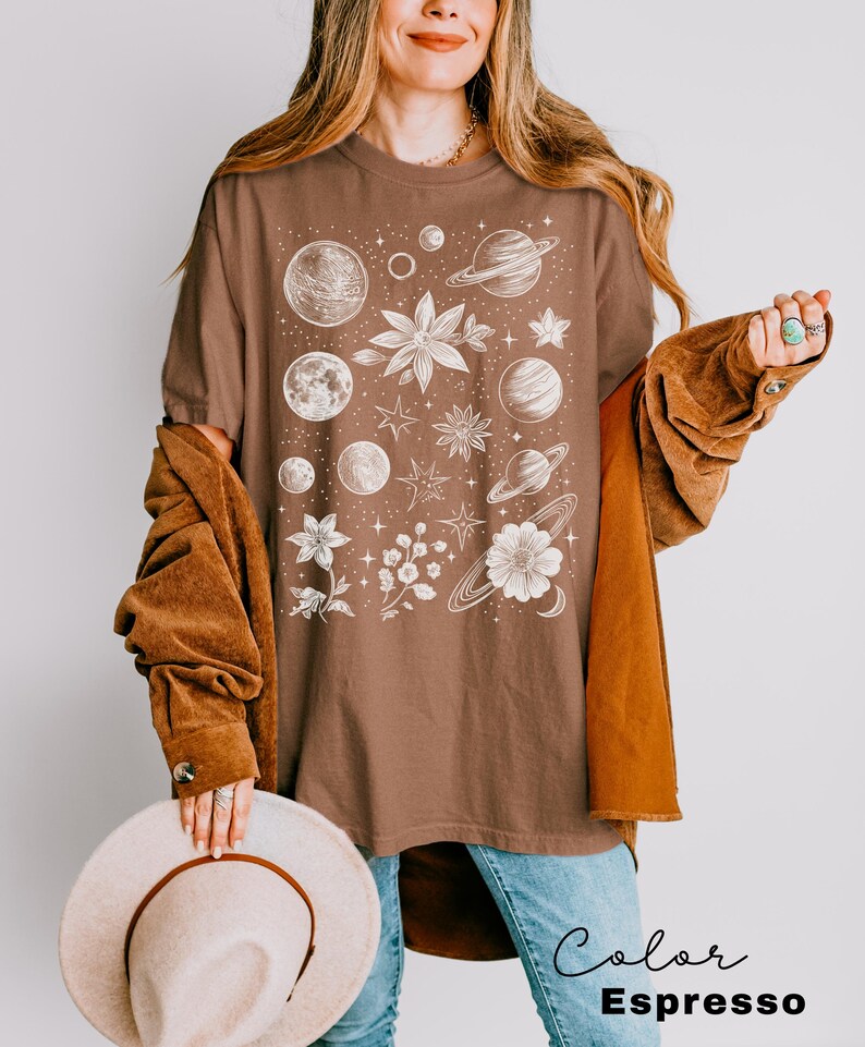 Puede incluir: Una camiseta marr&oacute;n con un estampado gr&aacute;fico blanco de planetas, estrellas y flores. El texto "Color Espresso" est&aacute; impreso debajo de la imagen.