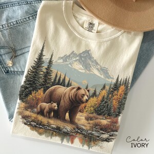 May include: Ivory-colored t-shirt featuring a detailed illustration of a brown bear and cub in a forest setting with a mountain backdrop. The design includes autumn foliage and a reflective lake. The shirt is paired with blue jeans and a tan hat.