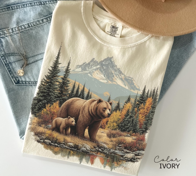 May include: Ivory-colored t-shirt featuring a detailed illustration of a brown bear and cub in a forest setting with a mountain backdrop. The design includes autumn foliage and a reflective lake. The shirt is paired with blue jeans and a tan hat.