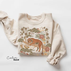 May include: Sand-colored sweatshirt featuring a detailed illustration of a tiger resting on a tree branch, surrounded by floral and cloud motifs. The design incorporates shades of orange, brown, green, and cream. The word "Color" and "Sand" are written in script.