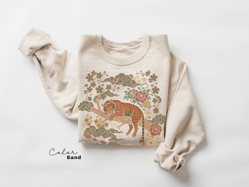 May include: Sand-colored sweatshirt featuring a detailed illustration of a tiger resting on a tree branch, surrounded by floral and cloud motifs. The design incorporates shades of orange, brown, green, and cream. The word "Color" and "Sand" are written in script.