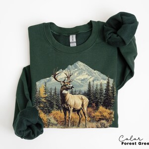 May include: Forest green sweatshirt featuring a detailed illustration of a deer with large antlers standing in front of a mountain range and evergreen trees. The text "Color Forest Green" is at the bottom right.
