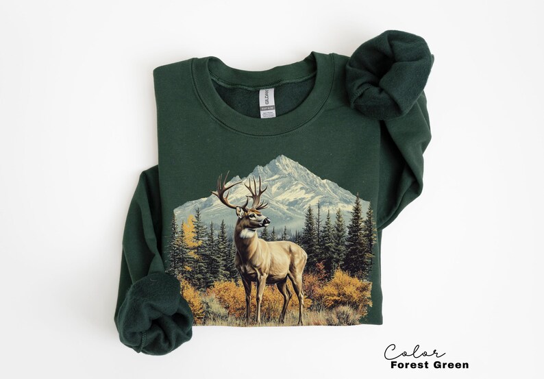 May include: Forest green sweatshirt featuring a detailed illustration of a deer with large antlers standing in front of a mountain range and evergreen trees. The text "Color Forest Green" is at the bottom right.