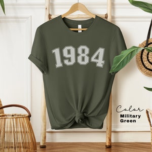 May include: A green t-shirt with the number 1984 printed in white on the front. The shirt is made of a soft, comfortable material and is perfect for everyday wear.