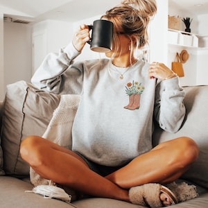 Puede incluir: Sudadera gris claro con un dise&ntilde;o floral de bota de vaquero. La bota est&aacute; llena de flores coloridas. El texto "Color Ash" est&aacute; en la parte inferior de la imagen. Se sostiene una taza negra.