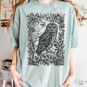 May include: A light teal t-shirt featuring a detailed black and white illustration of an owl within a floral frame. The owl is the focal point, surrounded by intricate botanical elements and butterflies. The shirt has short sleeves and a relaxed fit.