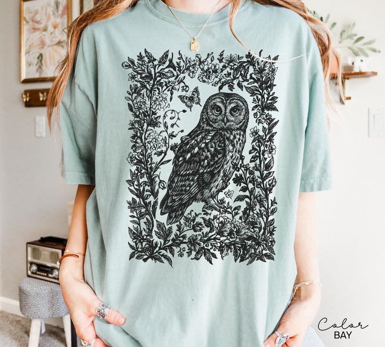 May include: A light teal t-shirt featuring a detailed black and white illustration of an owl within a floral frame. The owl is the focal point, surrounded by intricate botanical elements and butterflies. The shirt has short sleeves and a relaxed fit.
