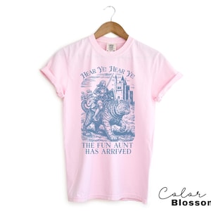May include: A light pink t-shirt with a blue graphic of a person riding a tiger, a castle, and the text "Hear Ye! Hear Ye! The Fun Aunt Has Arrived." The shirt is hanging on a wooden hanger.