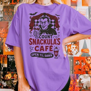 May include: A violet-colored t-shirt featuring a graphic of Count Snackula holding a milkshake. The design includes bats, a checkered curtain, and the text "COUNT SNACKULA'S CAFE OPEN TIL DAWN". A vintage-style ice cream cone is also shown.
