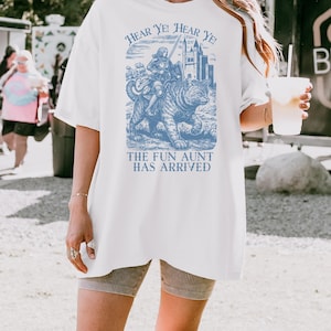May include: White t-shirt with a blue graphic featuring a knight riding a cat, accompanied by the text "HEAR YE! HEAR YE!" and "THE FUN AUNT HAS ARRIVED." The shirt has short sleeves and a relaxed fit.