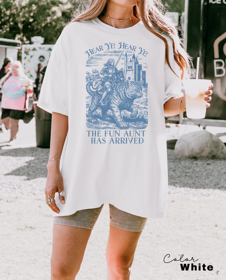 May include: White t-shirt with a blue graphic featuring a knight riding a cat, accompanied by the text "HEAR YE! HEAR YE!" and "THE FUN AUNT HAS ARRIVED." The shirt has short sleeves and a relaxed fit.