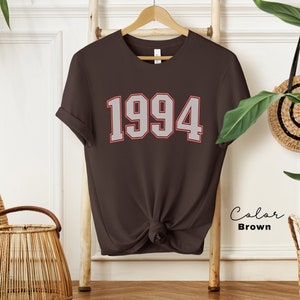 May include: A brown t-shirt with the year 1994 printed in white with a red outline. The shirt is on a hanger and is tied at the bottom.
