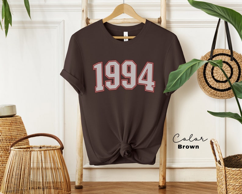 May include: A brown t-shirt with the year 1994 printed in white with a red outline. The shirt is on a hanger and is tied at the bottom.