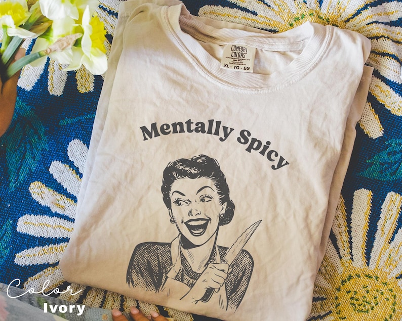 Puede incluir: Camiseta de color marfil con una ilustraci&oacute;n retro de una mujer sosteniendo un cuchillo y el texto "Mentally Spicy".