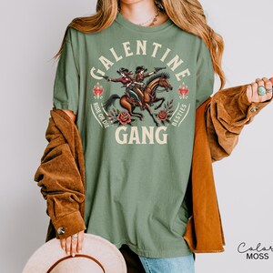 May include: Sage green t-shirt with a graphic of two cowgirls riding a horse, with the words "GALENTINE GANG" and "Ride or Die Besties". The shirt has a vintage, western aesthetic.