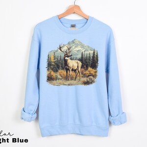 May include: Light blue sweatshirt featuring a detailed graphic of a deer with large antlers standing in a mountain landscape. The design includes trees and a mountain range in the background. The text "Color Light Blue" is at the bottom.