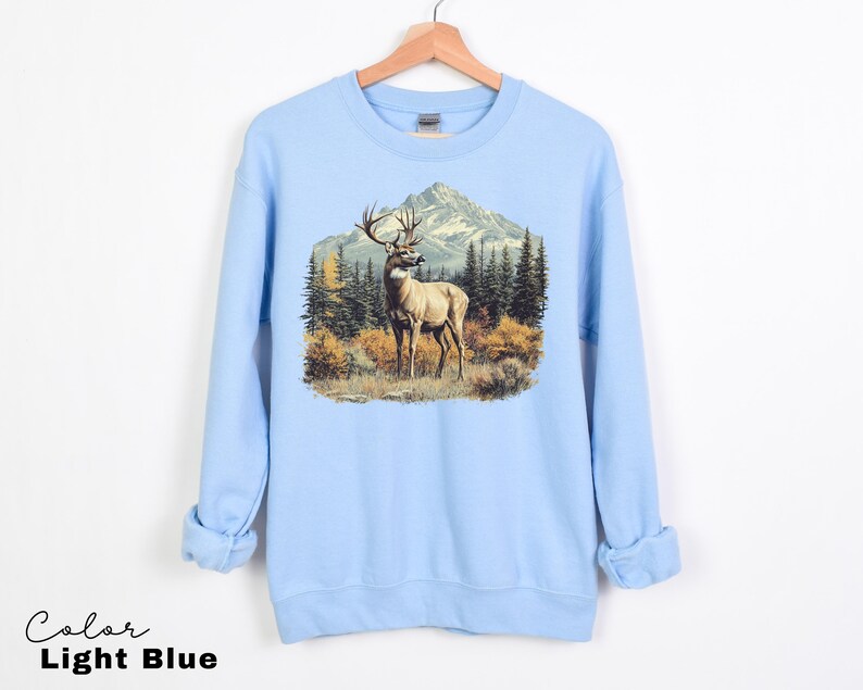May include: Light blue sweatshirt featuring a detailed graphic of a deer with large antlers standing in a mountain landscape. The design includes trees and a mountain range in the background. The text "Color Light Blue" is at the bottom.