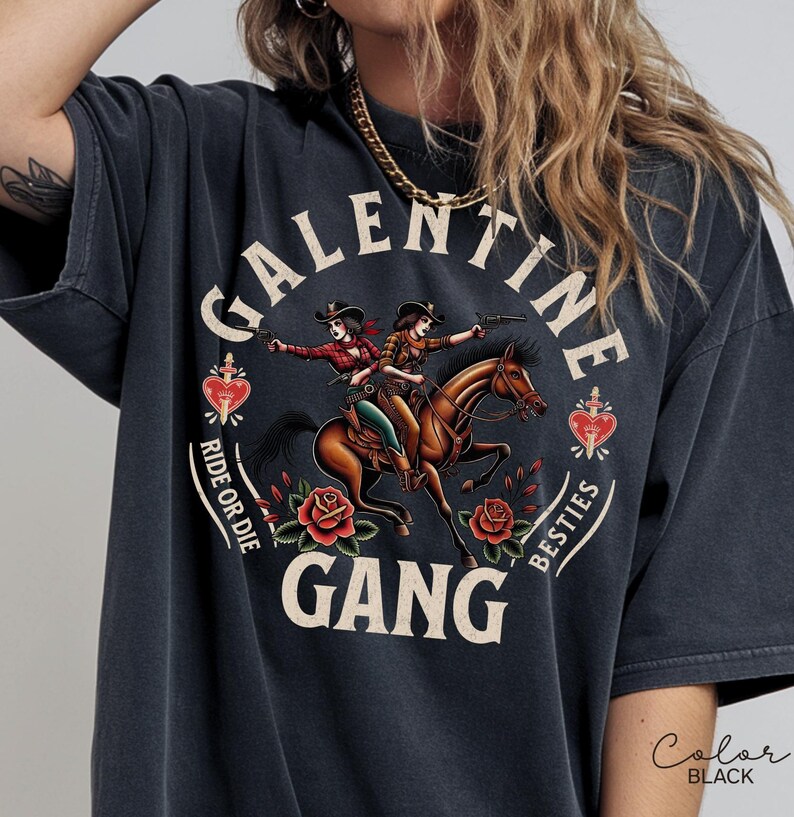May include: Black t-shirt with a graphic featuring two cowgirls riding a horse, guns drawn. The text reads "GALENTINE GANG" with "Ride or Die" and "Besties" around the image. Red roses and hearts complete the design.