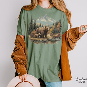 May include: Sage green t-shirt featuring a detailed graphic of a bear and cub in a forest scene with mountains and a lake. The design includes trees, a mountain range, and a setting sun. The text "Color MOSS" is in the lower right corner.