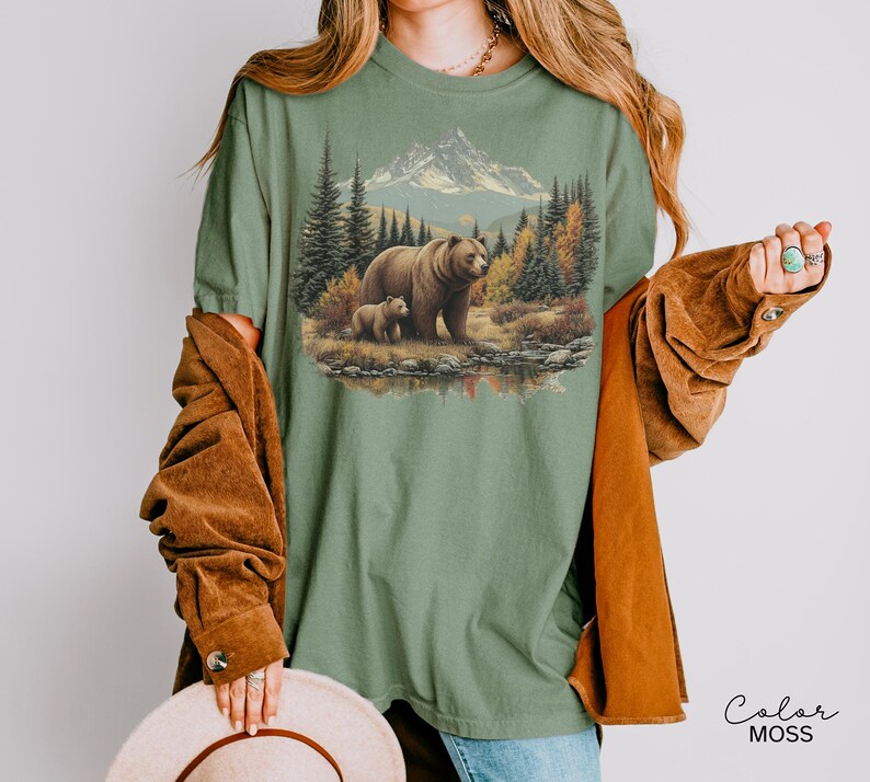 May include: Sage green t-shirt featuring a detailed graphic of a bear and cub in a forest scene with mountains and a lake. The design includes trees, a mountain range, and a setting sun. The text "Color MOSS" is in the lower right corner.