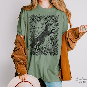 Pu&ograve; includere: T-shirt verde salvia con un'illustrazione in bianco e nero di una volpe che salta all'interno di una cornice floreale, con farfalle. La maglietta ha una vestibilit&agrave; ampia e maniche corte. Il testo "Color MOSS" &egrave; in basso a destra.