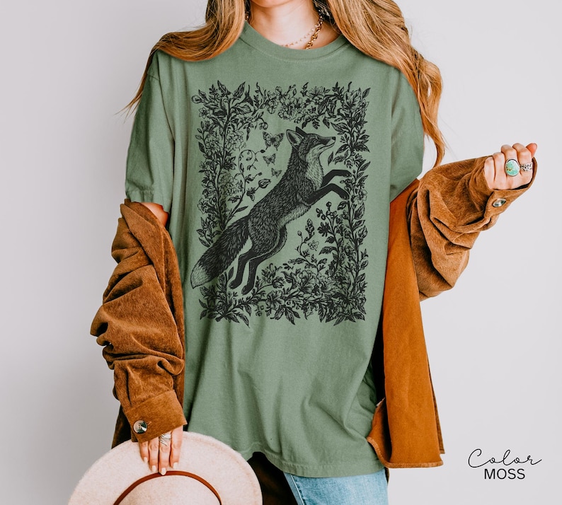 Pu&ograve; includere: T-shirt verde salvia con un'illustrazione in bianco e nero di una volpe che salta all'interno di una cornice floreale, con farfalle. La maglietta ha una vestibilit&agrave; ampia e maniche corte. Il testo "Color MOSS" &egrave; in basso a destra.