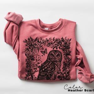May include: Heather Scarlet Red sweatshirt with a black graphic of an owl surrounded by floral elements and butterflies. The text "Color Heather Scarlet Red" is at the bottom right.