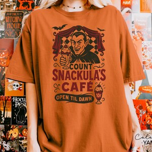 May include: An orange t-shirt featuring a graphic of Count Snackula holding a sundae, with the text "COUNT SNACKULA'S CAFE OPEN TIL DAWN". The design includes bats and a vintage cafe sign. A Halloween-themed apparel item.