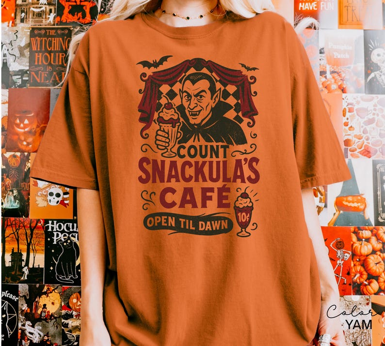 May include: An orange t-shirt featuring a graphic of Count Snackula holding a sundae, with the text "COUNT SNACKULA'S CAFE OPEN TIL DAWN". The design includes bats and a vintage cafe sign. A Halloween-themed apparel item.