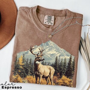 May include: Espresso-colored t-shirt featuring a mountain scene with a deer. The graphic includes a mountain range, evergreen trees, and autumn foliage. A silver necklace with a pendant rests on the shirt. The shirt has a Comfort Colors tag.