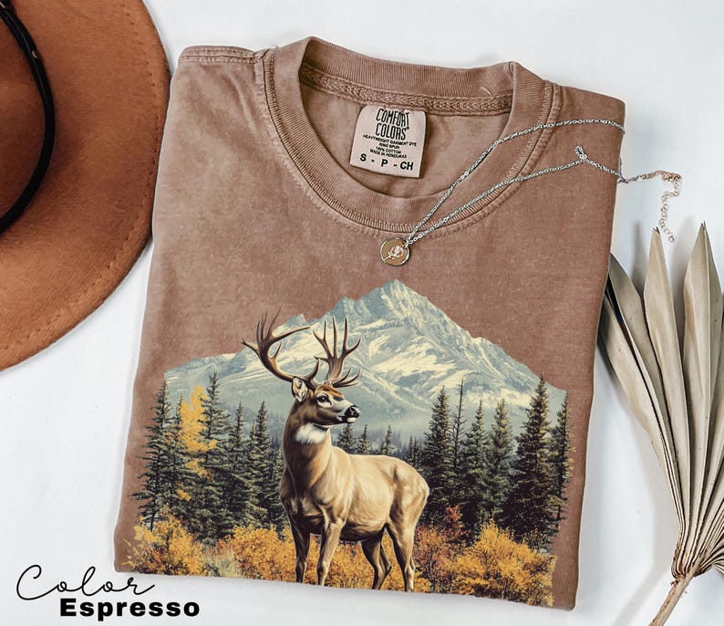 May include: Espresso-colored t-shirt featuring a mountain scene with a deer. The graphic includes a mountain range, evergreen trees, and autumn foliage. A silver necklace with a pendant rests on the shirt. The shirt has a Comfort Colors tag.