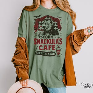 May include: Sage green t-shirt with a graphic of Count Snackula's Cafe. The design features a vampire holding a milkshake, bats, and the text "Count Snackula's Cafe Open Til Dawn".