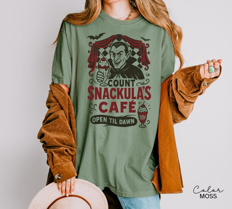 May include: Sage green t-shirt with a graphic of Count Snackula's Cafe. The design features a vampire holding a milkshake, bats, and the text "Count Snackula's Cafe Open Til Dawn".