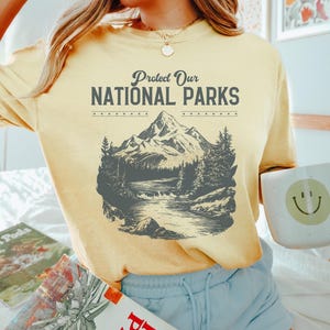May include: A yellow t-shirt with a vintage-style graphic of a mountain range and a river. The text "Protect Our National Parks" is printed on the shirt.