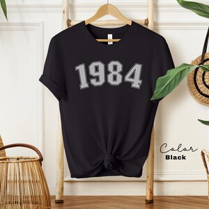 May include: Black t-shirt with the number 1984 printed in a gray, varsity-style font.