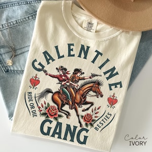 May include: Ivory-colored t-shirt with a graphic featuring two cowgirls riding a horse, guns drawn, and the text "Galentine Gang Besties Ride or Die." The design includes red roses and a heart.