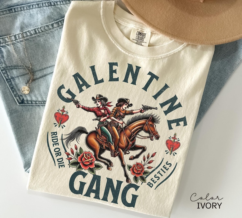 May include: Ivory-colored t-shirt with a graphic featuring two cowgirls riding a horse, guns drawn, and the text "Galentine Gang Besties Ride or Die." The design includes red roses and a heart.
