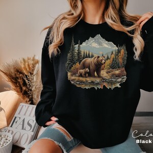May include: Black sweatshirt featuring a detailed illustration of a bear and cub in a forest scene with a mountain backdrop. The design includes trees, a lake, and autumn colors. The word "Color" and "Black" are written on the bottom right.