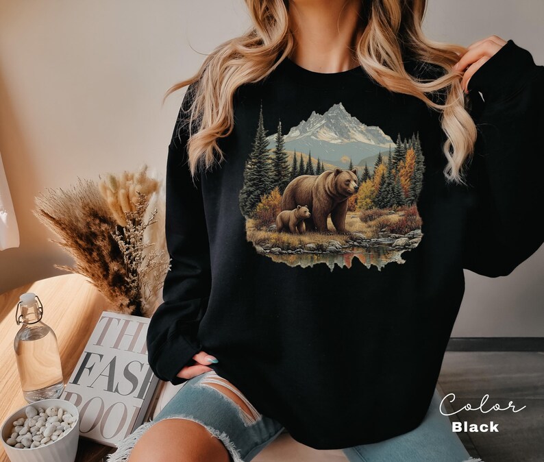 May include: Black sweatshirt featuring a detailed illustration of a bear and cub in a forest scene with a mountain backdrop. The design includes trees, a lake, and autumn colors. The word "Color" and "Black" are written on the bottom right.