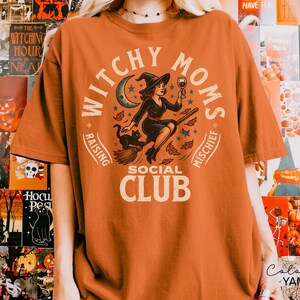Puede incluir: Camiseta naranja con un dise&ntilde;o de una bruja en una escoba, un gato negro y las palabras "Witchy Moms Social Club". El dise&ntilde;o incluye una luna creciente, estrellas y hojas de oto&ntilde;o. La camiseta es un art&iacute;culo tem&aacute;tico de Halloween.